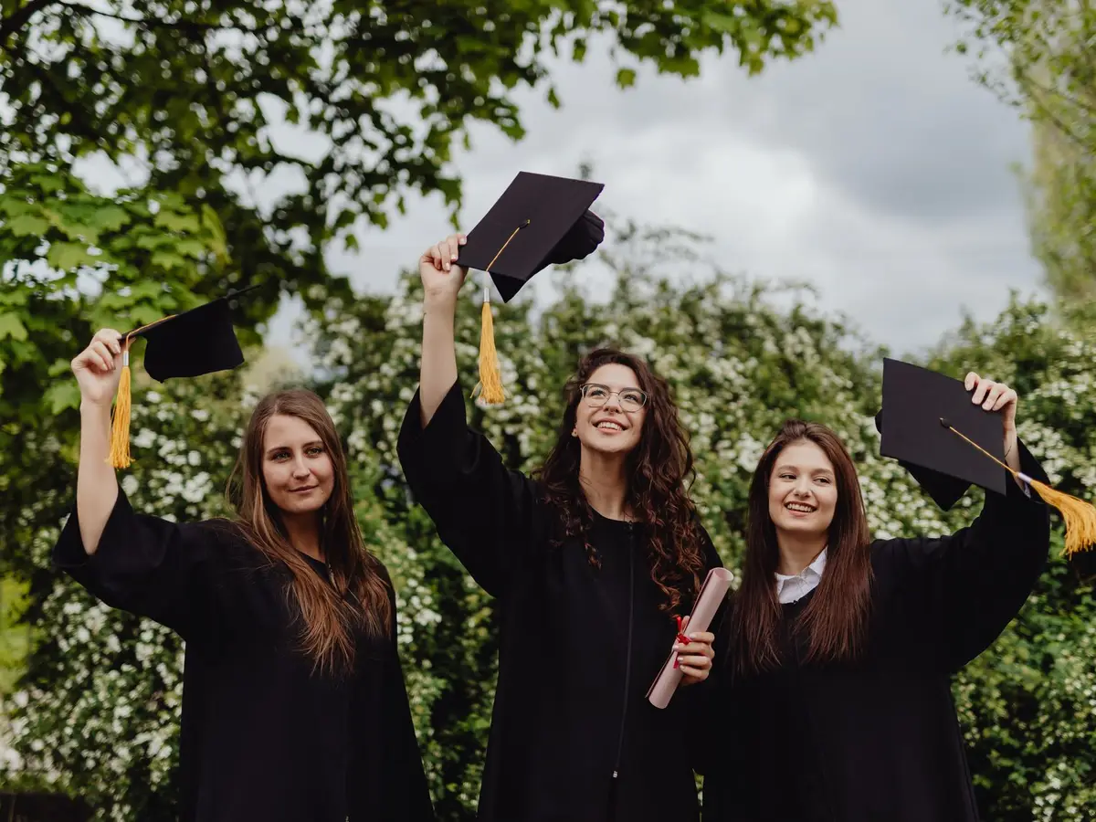 Three graduates in black gowns holding their caps up outdoors