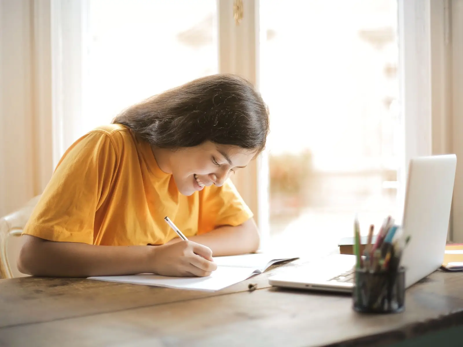 A student smiling while writing a practice essay, with a laptop open on the desk