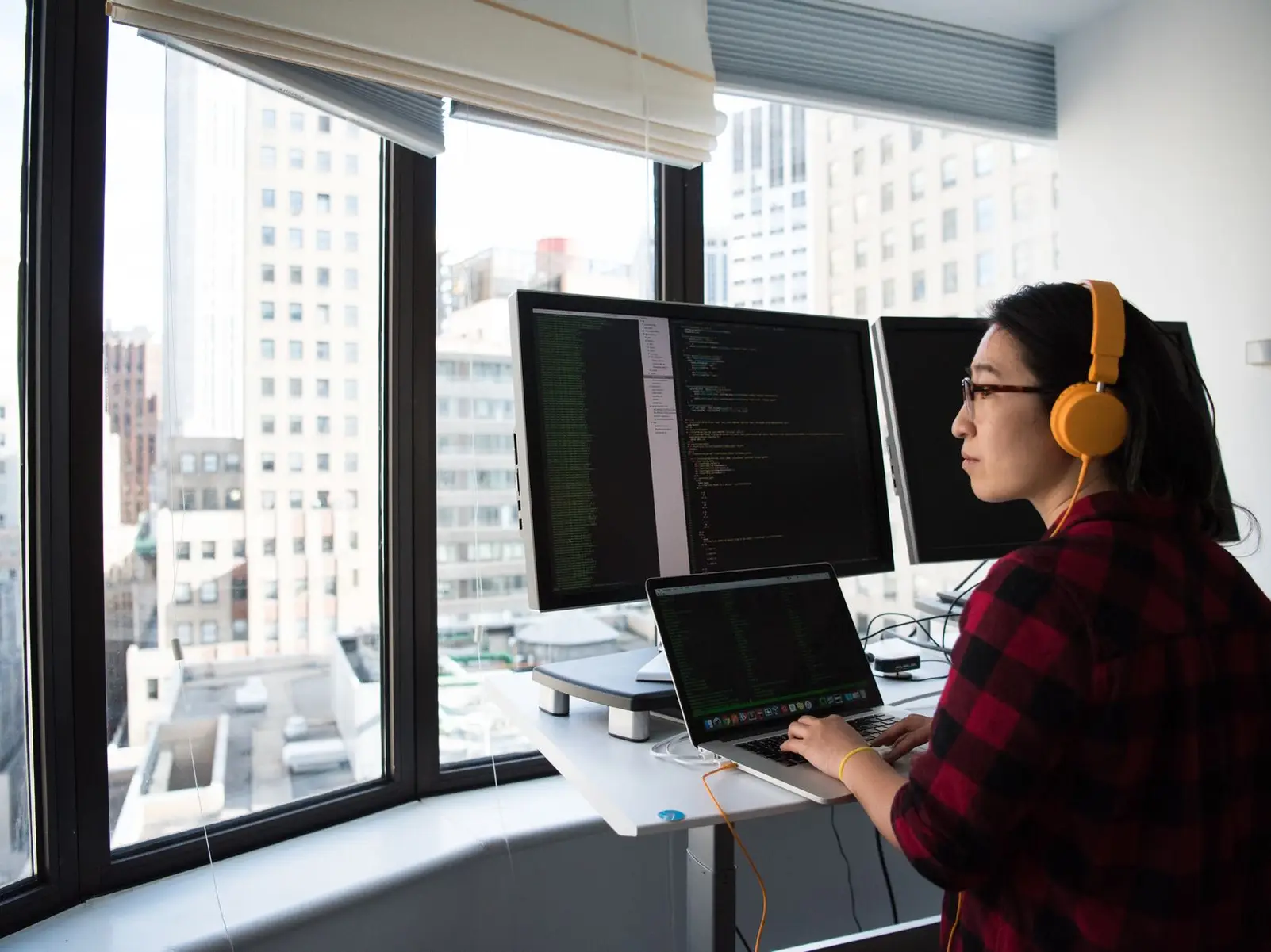 A developer in headphones writing code at a standing desk, city towers outside the window