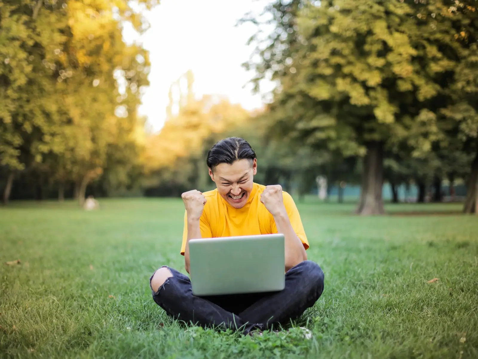 A student sitting on the grass in a park, celebrating with both fists raised in front of a laptop