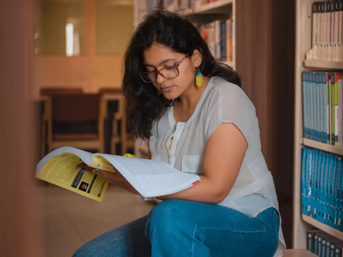A student reading a book in a library, shelves of books behind her