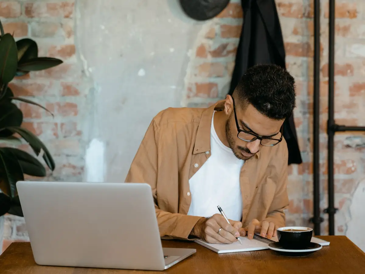 A young man writing in a notebook at a desk beside his laptop