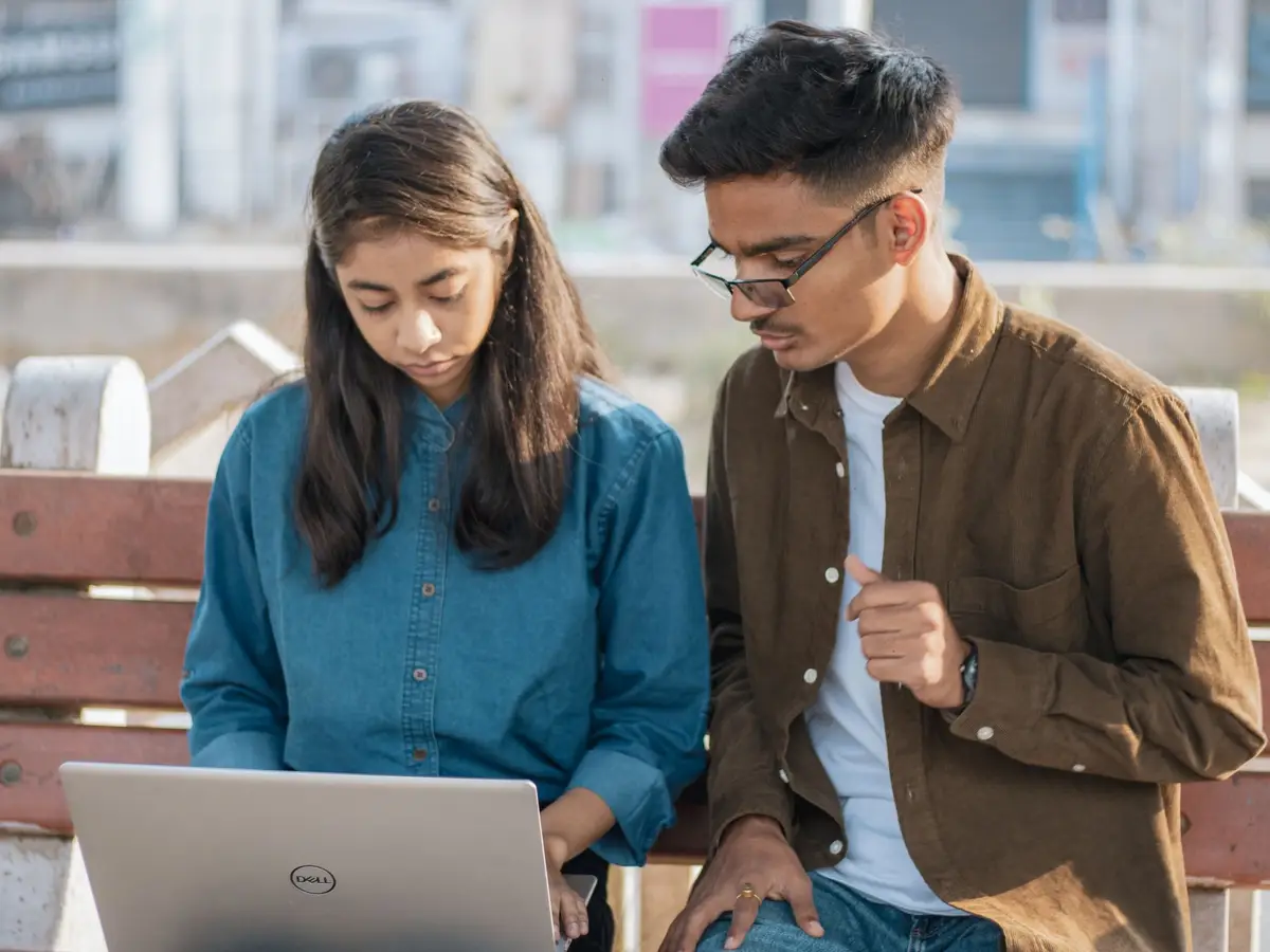 Two young students checking an application on a laptop on a park bench