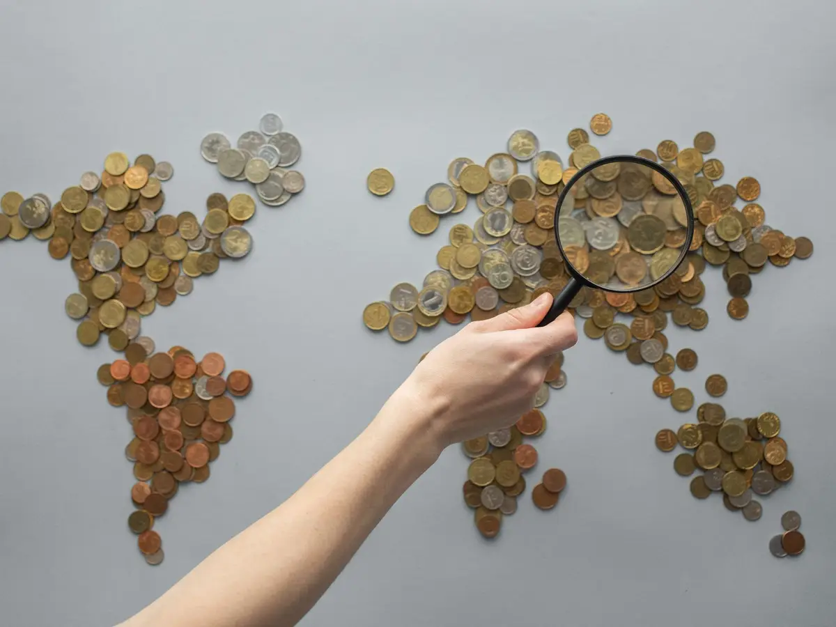 A world map made of coins, with a hand holding a magnifying glass over it
